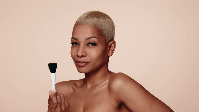 Self-assured young Hispanic woman with short hair showcasing a makeup brush, posing against a nude tone background.