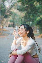 A young woman enjoys a peaceful moment, holding a clear water bottle with a gentle smile, as she takes a hydration break during an outdoor workout, wearing a smartwatch, wireless earbuds.