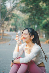 Fototapeta premium A young woman enjoys a peaceful moment, holding a clear water bottle with a gentle smile, as she takes a hydration break during an outdoor workout, wearing a smartwatch, wireless earbuds.