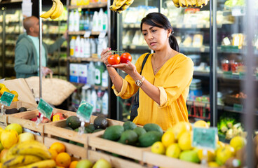 Hispanic female shopper looking for fresh organic tomatoes in greengrocery department of supermarket