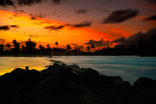 San Juan Puerto Rico Skyline at Stormy Sunset with dramatic clouds, orange sky, Palm trees, and sea waves over the volcanic rocks on La Poza del Escambr&oacute;n Beach and Escambron Beach