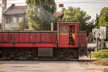 Red locomotive, a Kleinlokomotive, an old diesel shunting locomotive passing a level crossing in Smederevo train station, preparing a cargo freight train, old and decaying, fuming and smoking.