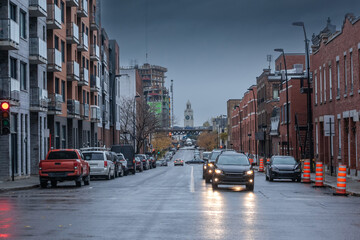 Selective blur on cars driving at dusk in Saint henri district of Montreal residential North American street with typical red brick buildings cars driving & cars parked. Atwater Market Tower is visibl - obrazy, fototapety, plakaty