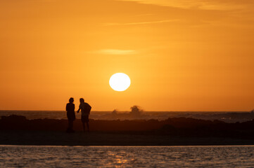 Man and woman staying and looking at sunset on ocean beach, orange sky, silhouettes of people on vacation