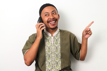 Indonesian Muslim man in koko and peci points to the side at copy space while talking on his phone,...