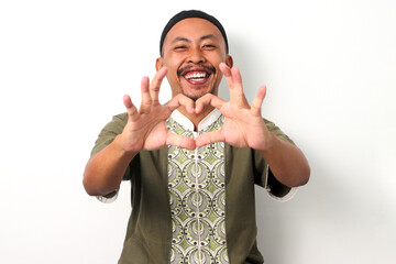 Smiling Indonesian Muslim man in koko shirt and peci makes a heart gesture with his hands,...