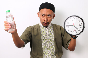 An Indonesian Muslim man in koko and peci holds a mineral water bottle and a clock, looking...