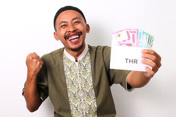 An excited Indonesian Muslim man in koko and peci holds a white envelope labeled THR, filled with money, representing his Religious Holiday Allowance. Isolated on a white background