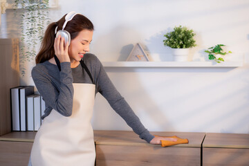 Excited young asian woman with headphones singing and dancing while dusting a wooden cabinet in...