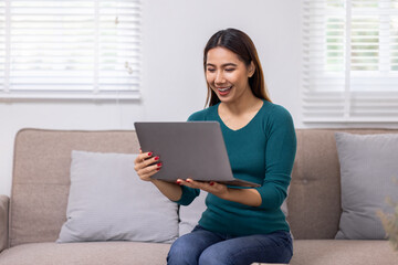 Happy young asian woman relax on comfortable couch at home texting messaging on laptop smartphone, smiling girl use cellphone chatting, browse wireless internet on gadget, work online from home