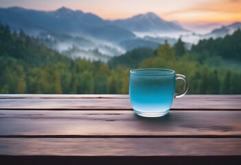 Transparent mug of tea on a wooden table with a serene mountain landscape and misty sunrise in the background. - obrazy, fototapety, plakaty
