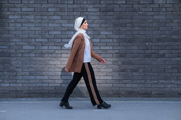 A young woman dressed in a hijab and a business suit walks along a brick wall. 