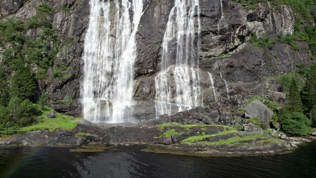 Camera pushes in on the bottom of a large waterfall, Laukelandsfossen, in Norway. Water crashes into a small pool at the bottom and spray from the water creates rainbows in the sunlight.
