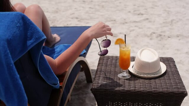 Hand of woman with sunglasses enjoying tanning in sunbed on sandy ocean beach, rear view. Cocktail orange fresh and sun hat near on wicker table. Tourism travel summer holidays vacations concept.