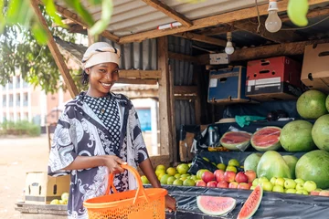 Fototapeten smiling woman holding shopping basket standing beside a farmers fruit shop © Boijonell