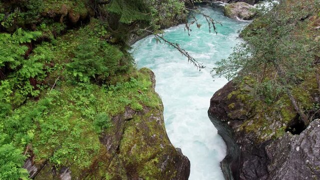 Crane shot, camera climbing from white water rushing between two moss covered rocks. Through the trees in the foreground, a large swirling waterfall is revealed in the background.