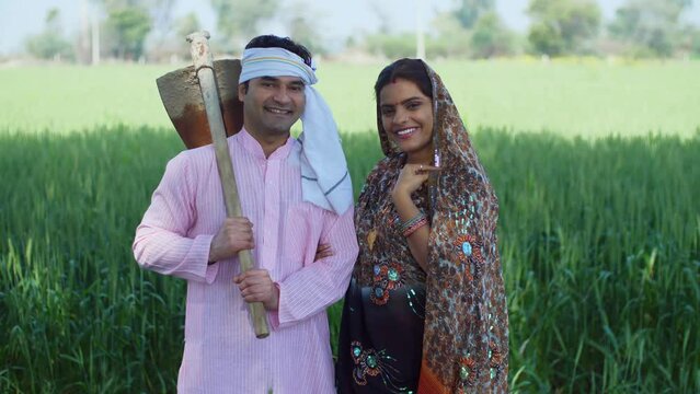 A desi Indian farmer with his wife posing happily for the camera - village life  rich village family  holding spade  farming tool. A rural couple in traditional wear - smiling  healthy green crops ...