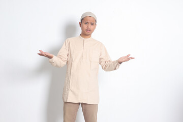 Portrait of confused Asian muslim man in koko shirt with skullcap spreading his hands sideways and...