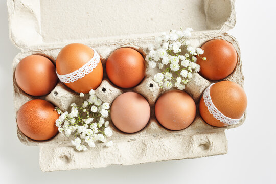 Easter eggs and white blooming flowers in carton package on light background. Easter holiday concept, top view. Close up chicken eggs decorated lace, symbol springtime holiday, spring celebration