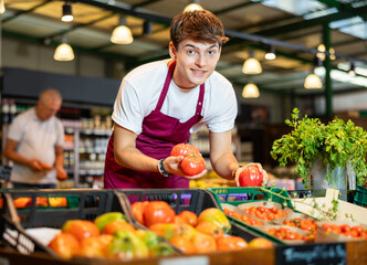 Cheerful young guy working in farm grocery store arranging ripe red tomatoes on counter with fresh vegetables and fruits