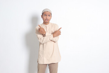 Portrait of excited Asian muslim man in koko shirt with skullcap pointing sideways, making choice,...