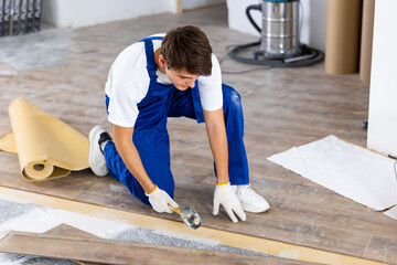 Skilled young carpenter using rubber mallet to lay click-lock laminate flooring during interior finishing work - 752014514