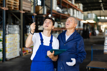 Elderly man and young guy warehouse workers in uniform discuss documents - 752014925