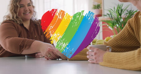 Image of rainbow heart over lesbian couple drinking coffee at home