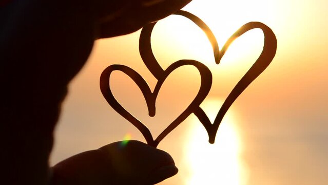 A woman holding in fingers a black outline in the shape of two hearts against the background of the sun at dawn and sunset on the sea. Concept love infatuation romantic Valentine's Day mood emotions
