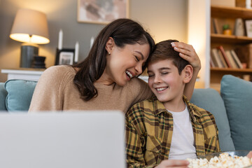 In their living room, a mom and her son have a fun movie night. With popcorn and a laptop, they watch a funny comedy. They laugh together at the jokes and funny scenes, enjoying each other's company.