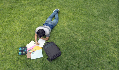 A male student lying on the grass writing in a notebook