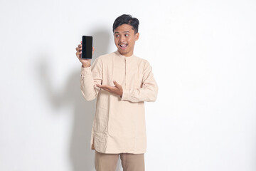 Portrait of young excited Asian muslim man in koko shirt showing blank screen mobile phone mockup while pointing and presenting product. Social media concept. Isolated image on white background