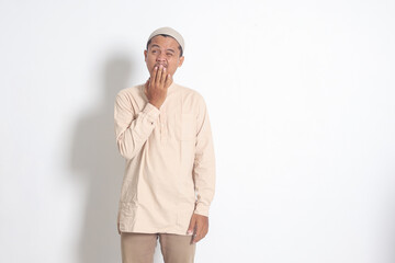 Portrait of overworked Asian muslim man in koko shirt with skullcap yawning with hand covering his...