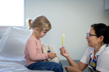 Smiling female nurse giving ice candy to girl sitting on bed in hospital