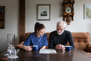 Senior man solving sudoku with female nurse having coffee while sitting on sofa in living room