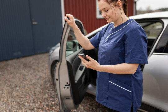 Female healthcare worker using mobile phone by car standing in back yard