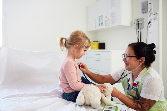 Smiling female pediatrician checking girl with stethoscope sitting on bed in hospital