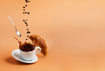 White coffee cup, plate and metal tea spoon falling on beige table background and coffee splashes because of hit, coffee beans levitate, fly and fall to cup, crispy fresh croissant. Breakfast concept