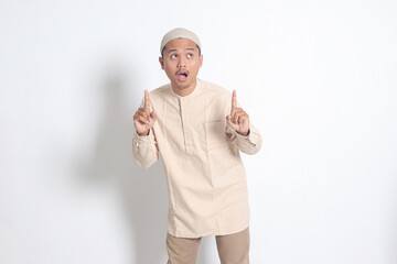 Portrait of shocked Asian muslim man in koko shirt with skullcap showing product and pointing with...