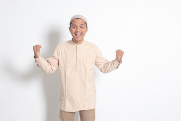 Portrait of religious Asian muslim man in koko shirt with skullcap raising his fist, celebrating...
