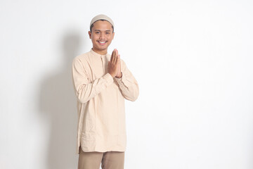Portrait of attractive Asian muslim man in koko shirt with skullcap showing apologize and welcome hand gesture. Apology during eid mubarak. Isolated image on white background - obrazy, fototapety, plakaty