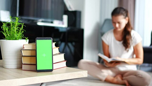 Digital detox concept photo. Smartphone with green chroma key screen and woman reading book in the background