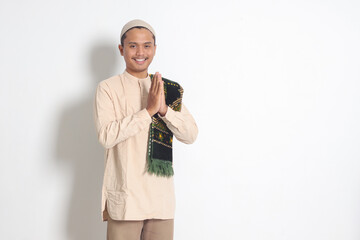 Portrait of attractive Asian muslim man in koko shirt with prayer mat showing apologize and welcome hand gesture. Apology during eid mubarak. Isolated image on white background