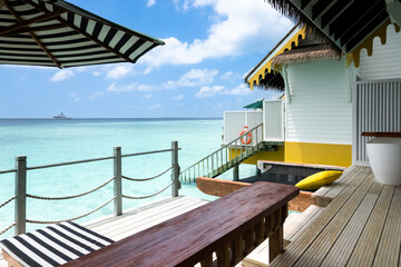 Beachfront deck of a tropical resort with striped awning, yellow house with a clear blue sky and seascape view of the turquoise ocean at Maldives island lifestyle