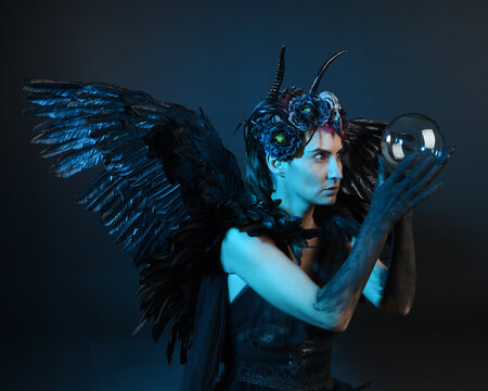 Close up portrait of female model wearing gothic horned headdress, halloween black dress, fantasy angel feather wings. Holding mystical crystal ball. dark studio background cinematic shadow silhouette