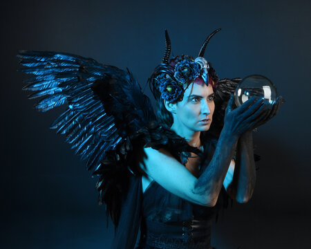 Close up portrait of female model wearing gothic horned headdress, halloween black dress, fantasy angel feather wings. Holding mystical crystal ball. dark studio background cinematic shadow silhouette