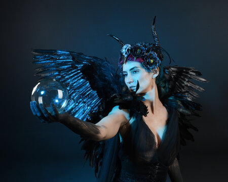 Close up portrait of female model wearing gothic horned headdress, halloween black dress, fantasy angel feather wings. Holding mystical crystal ball. dark studio background cinematic shadow silhouette