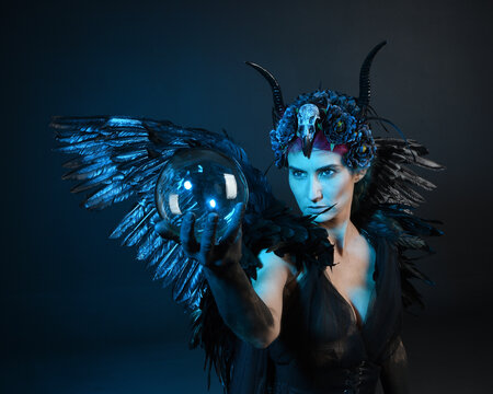 Close up portrait of female model wearing gothic horned headdress, halloween black dress, fantasy angel feather wings. Holding mystical crystal ball. dark studio background cinematic shadow silhouette