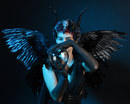 Close up portrait of female model wearing gothic horned headdress, halloween black dress, fantasy angel feather wings. Holding mystical crystal ball. dark studio background cinematic shadow silhouette