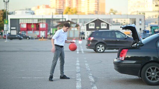 A young man in office clothes at the end of a work or school day plays with a basketball in the parking lot, throwing the ball into the trunk of a sedan